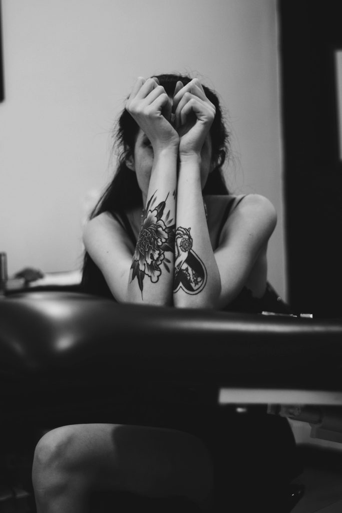 An artistic black and white portrait of a tattooed woman, arms raised, posing indoors.