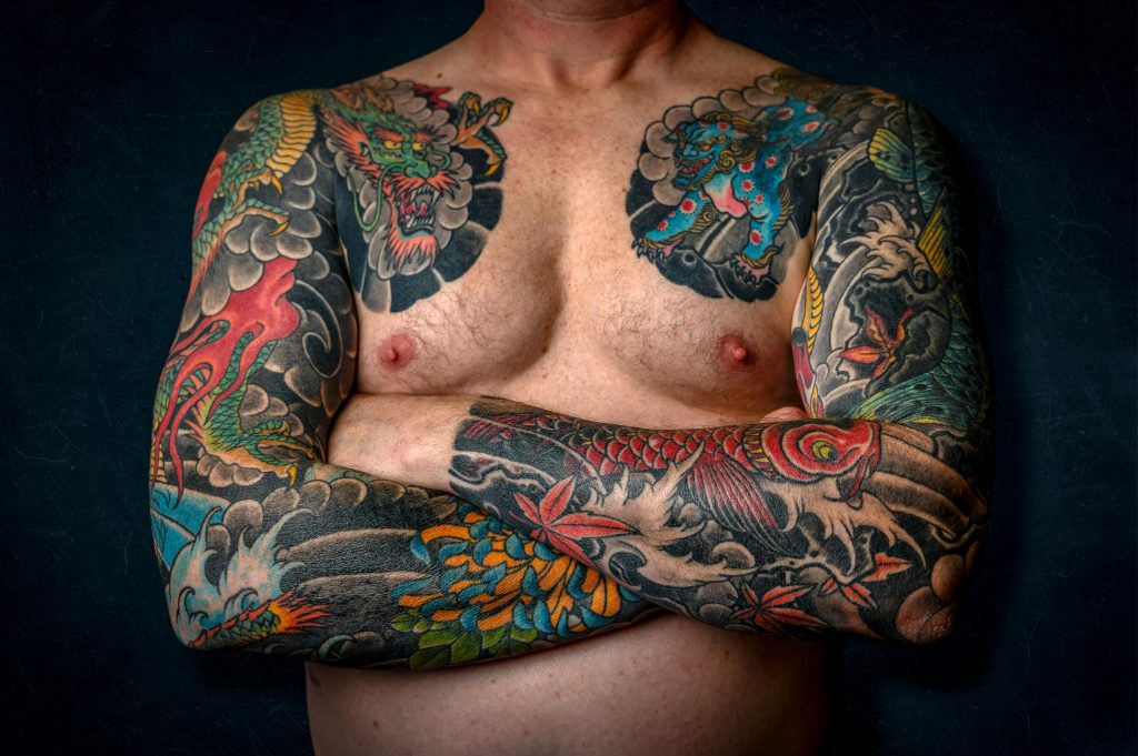 Close-up of a shirtless man with colorful sleeve tattoos and chest art.